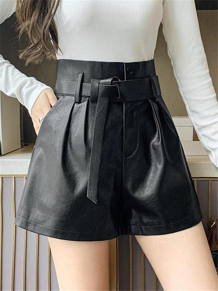 Jeune femme dont on ne voit que le haut des jambes porte un short noir plissé en simili cuir, une de ses mains est dans la poche, elle porte un pull blanc 